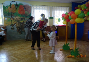 Pani Dyrektor przy bramce z balonami pasuje dziewczynkę czerwonym ołówkiem na przedszkolaka.